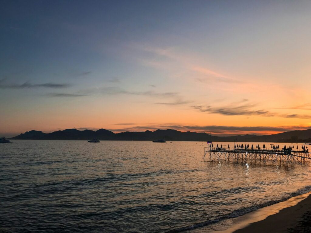 Solnedgang over roligt hav med udsigt til badebro med parasoller, skibe på vandet og bjerge i baggrunden.